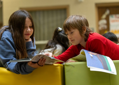 Photo of two children reading books together, the child on the left, with long hair and a blue sweatshirt, is holding a book. This child on the right, wearing a bright red shirt, is pointing at the book the child on the left is holding.